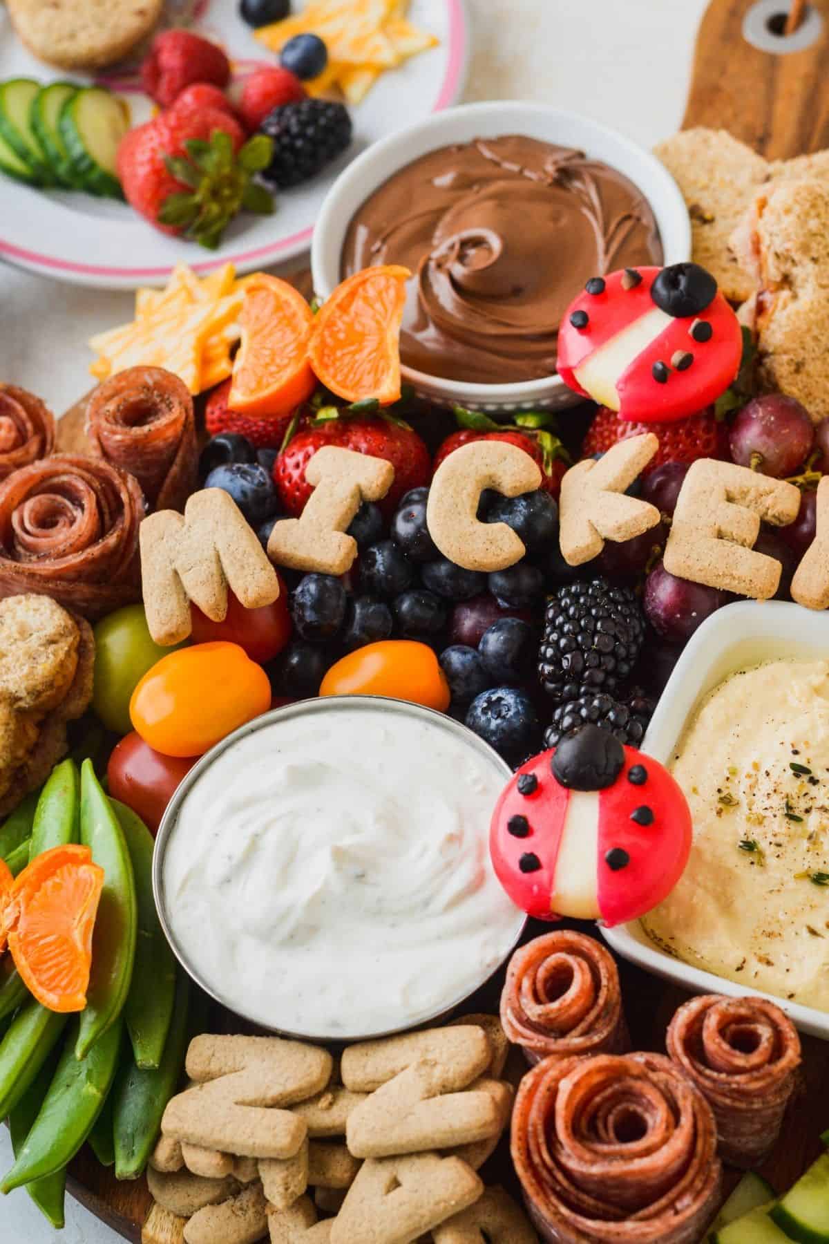 Charcuterie board filled with kid friendly foods like cinnamon alphabet cookies, fresh berries and Babybel ladybugs.