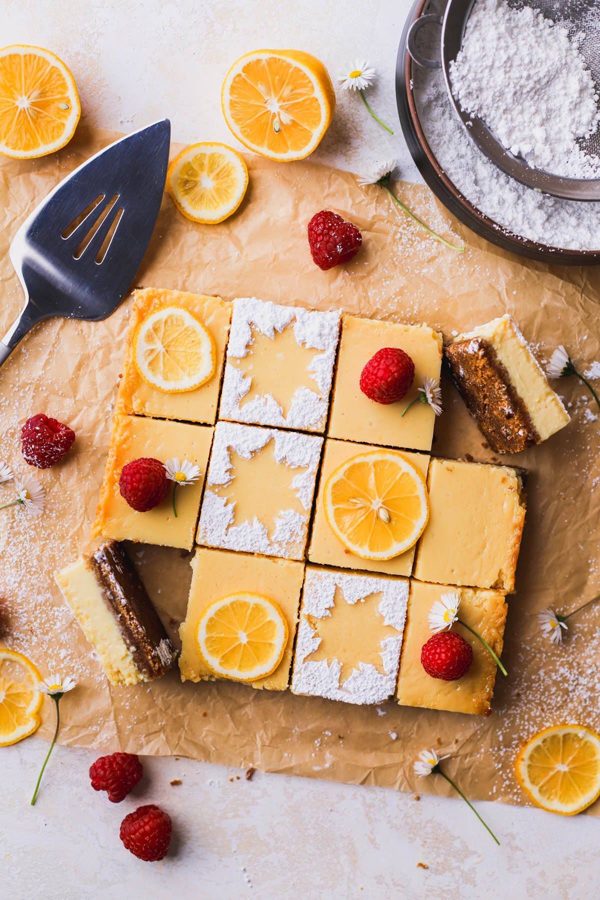 Lemon bars with graham cracker crust are dusted with powdered sugar and garnished with lemon slices and raspberries.