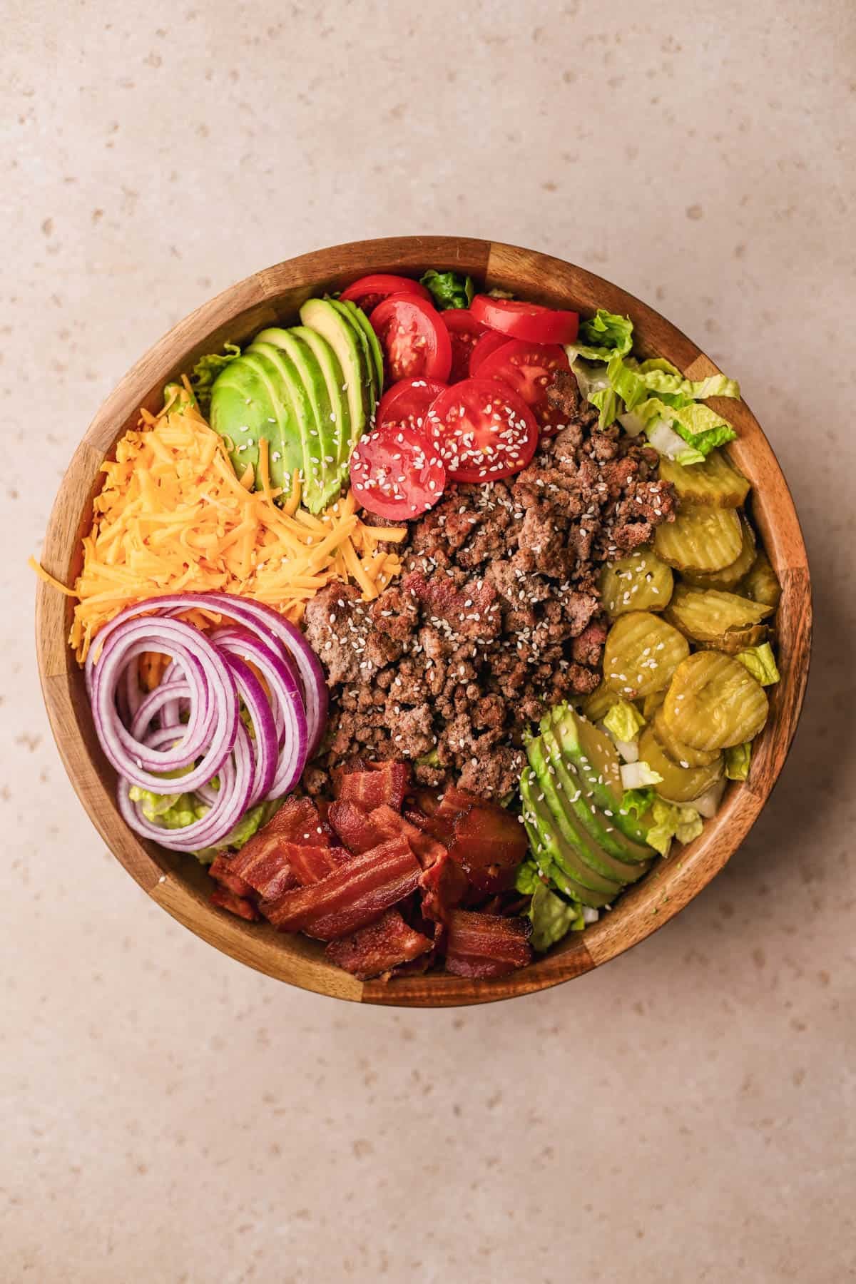Chopped romaine lettuce, sliced avocado, cheddar cheese, red onions, bacon, tomatoes, pickles, seared ground beef and white sesame seeds.