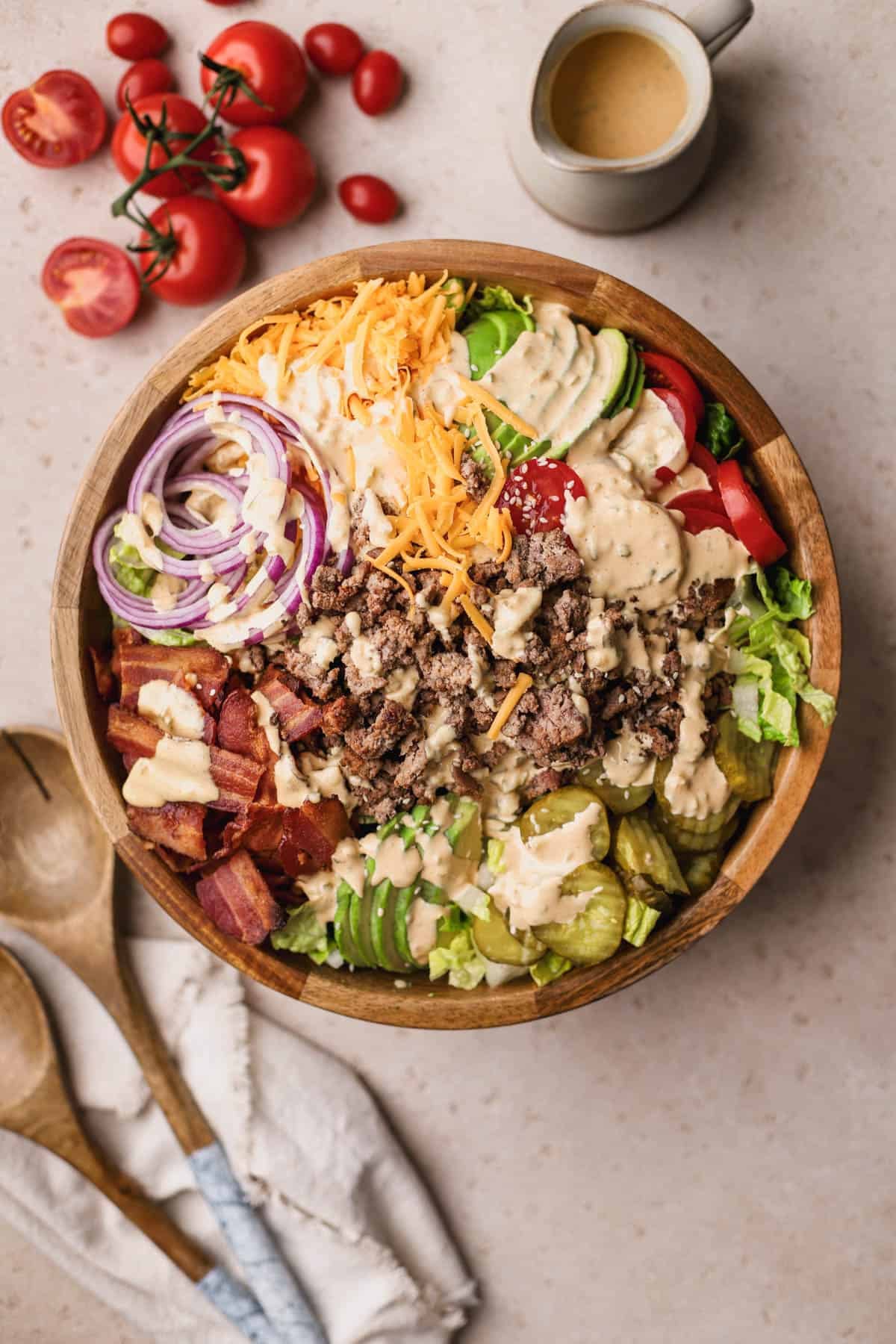 Deconstructed burger bowl is made with romaine lettuce, ground beef, tomatoes, red onions, pickles, sliced avocado, cheddar cheese, bacon and topped with special burger sauce and white sesame seeds.
