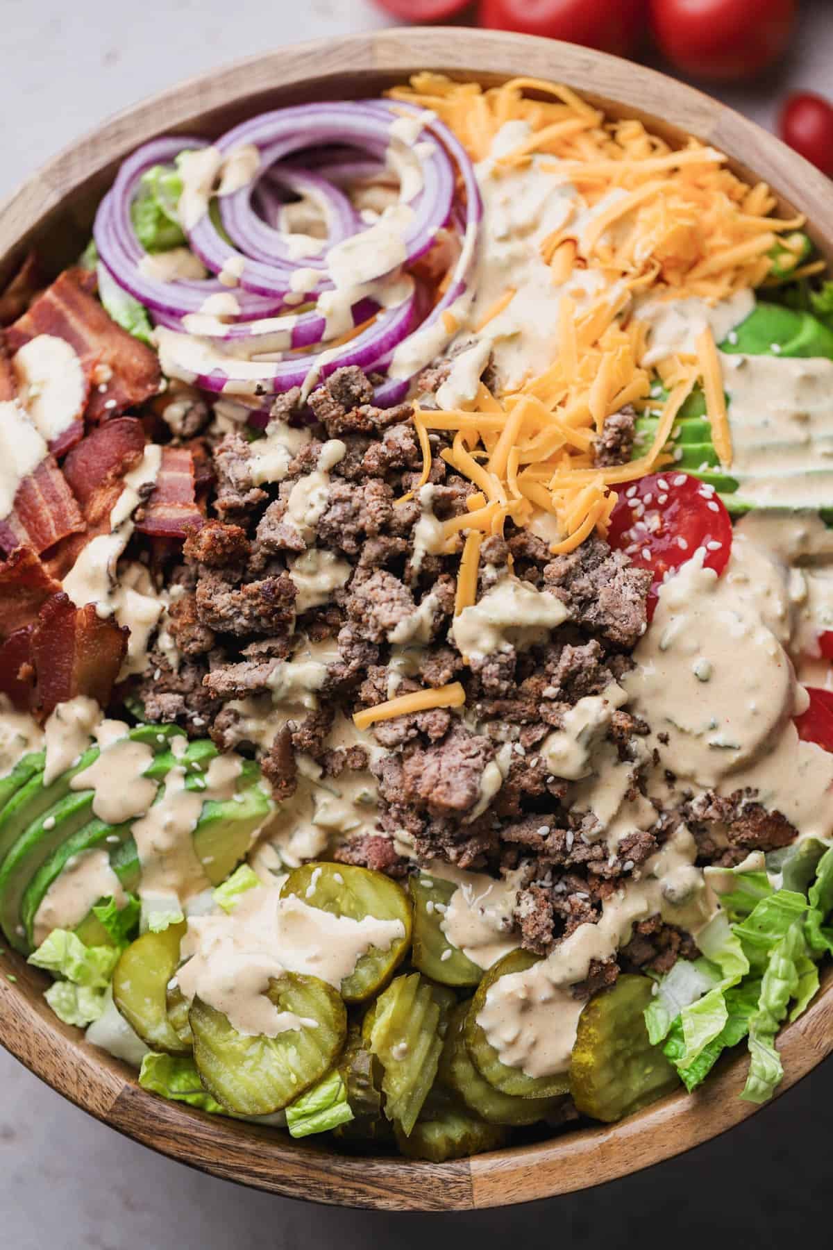 Deconstructed burger bowl is made with romaine lettuce, ground beef, tomatoes, red onions, pickles, sliced avocado, cheddar cheese, bacon and topped with special burger sauce and white sesame seeds.