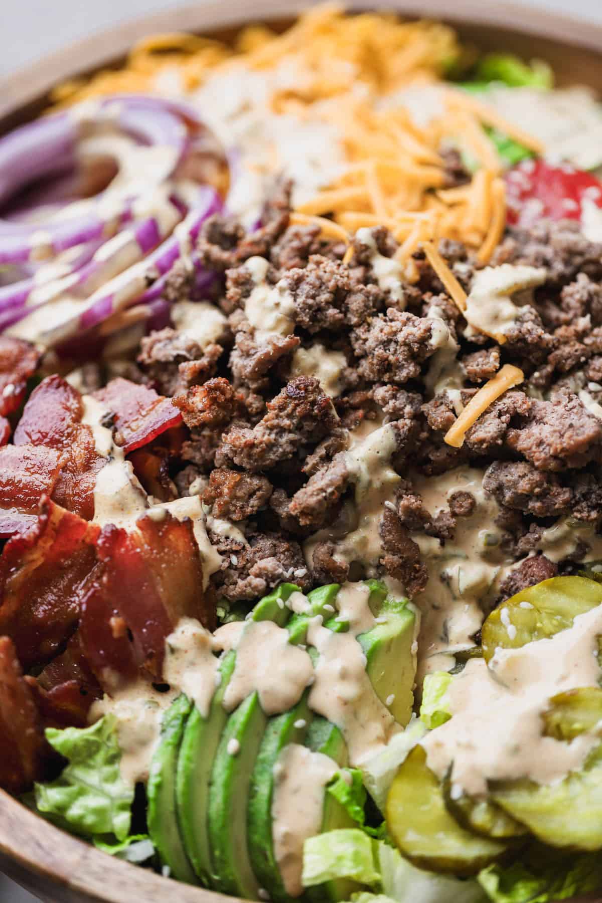 Deconstructed burger bowl is made with romaine lettuce, ground beef, tomatoes, red onions, pickles, sliced avocado, cheddar cheese, bacon and topped with special burger sauce and white sesame seeds.
