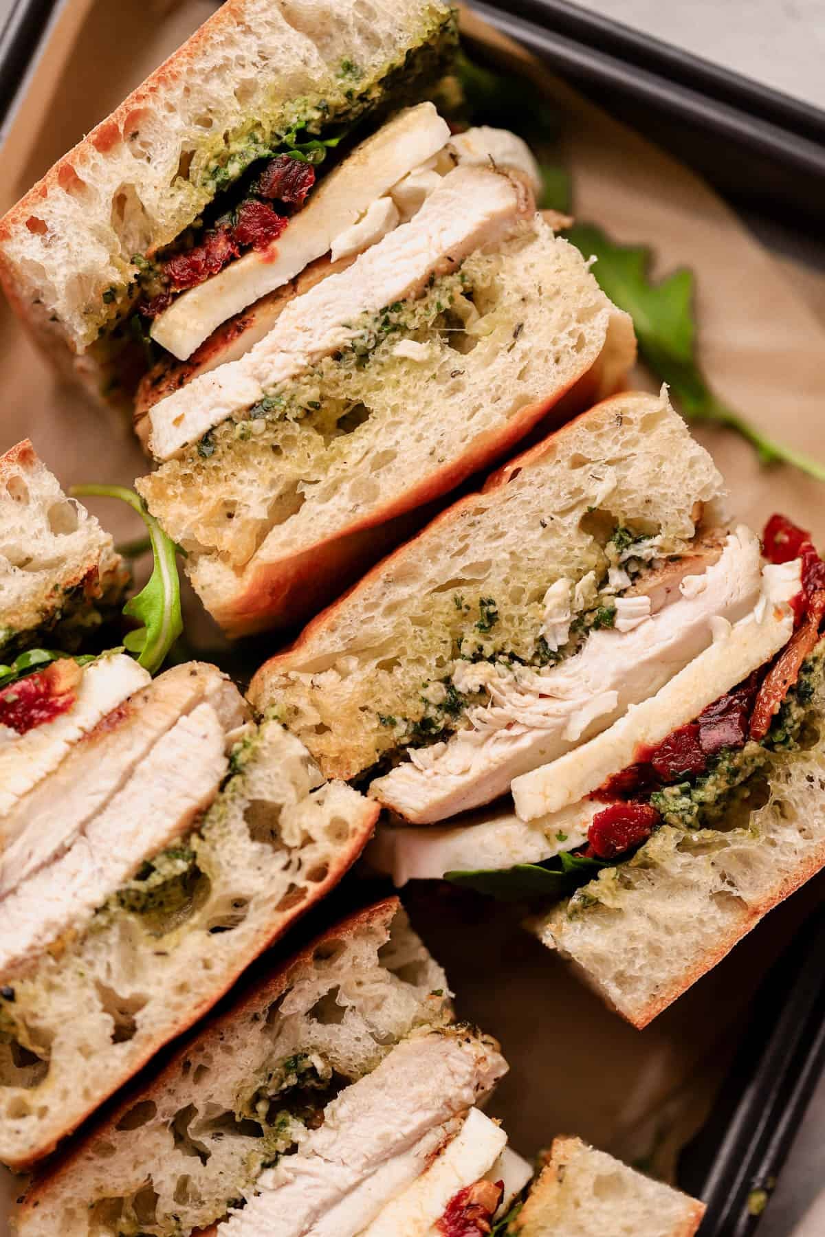 A Sun-Dried Tomato Pesto Chicken Sandwich starts with juicy chicken, marinated in Italian seasoning and olive oil, then it gets stacked on soft herb focaccia with a generous smear of homemade pesto, then layered with chewy sun-dried tomatoes, creamy fresh mozzarella, and a handful of peppery arugula.