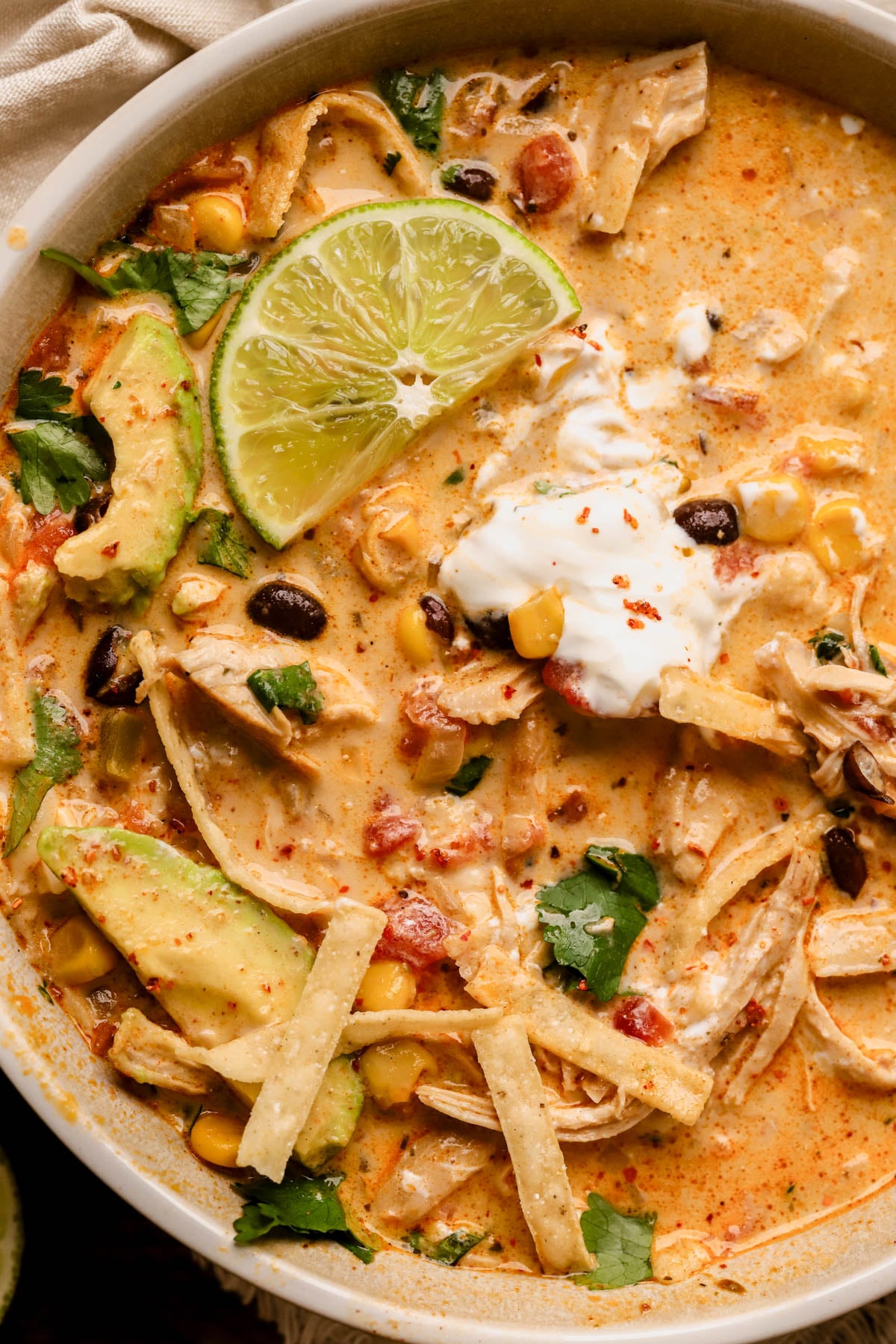 Close up of creamy chicken taco soup with avocado, lime, cilantro, sour cream, black beans, shredded chicken, crispy tortilla strips, and corn. 